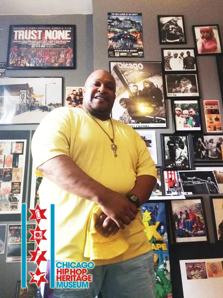 A man in a yellow shirt in front of a wall of framed photos and flyers.